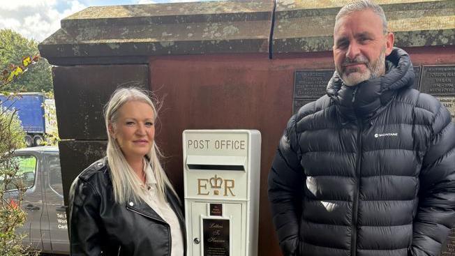 Michelle Milton, with long blonde hair, wearing a black leather jacket, stands next to the left of a white post office post box on a wall. To the right of it is Stephen Milton, with short grey hair, wearing a black puffer coat jacket. 