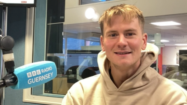 A man in a pale hoodie in a radio studio with a microphone with the words Radio Guernsey.