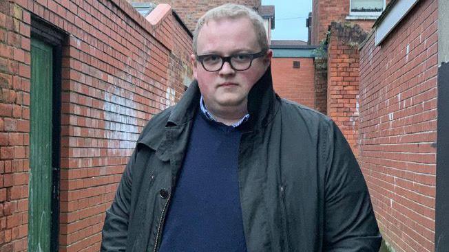 Séamas de Fáoite stands outside in an alley, He wears a blue jumper, blue shirt and grey jacket.