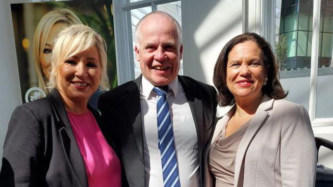 Sinn Féin candidate Gerard Magee wearing a dark suit, white shirt and blue and white striped tie, smiling along with deputy leader Michelle O'Neill wearing a pink top and dark jacket, and party leader Mary Lou McDonald, who is wearing a cream coloured jacket and top.