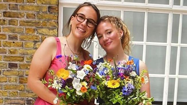 Jesse with her wife Faith seen holding flowers on their wedding day. Jesse, left, is wearing a pink dress, while Faith is in blue.