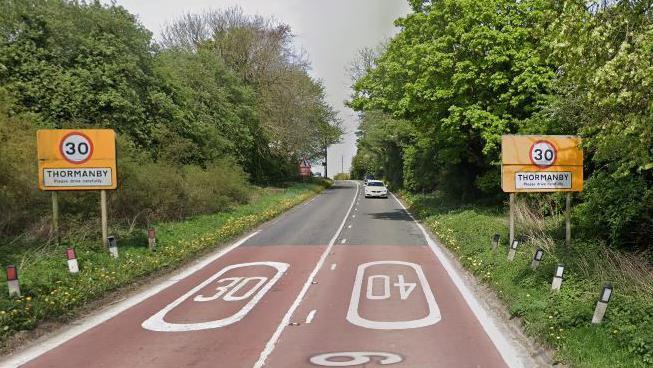 The A19 as it enters Thormanby with 30mph signs each side of the road and speed limit markers in white on the road and a Thormanby village sign post.