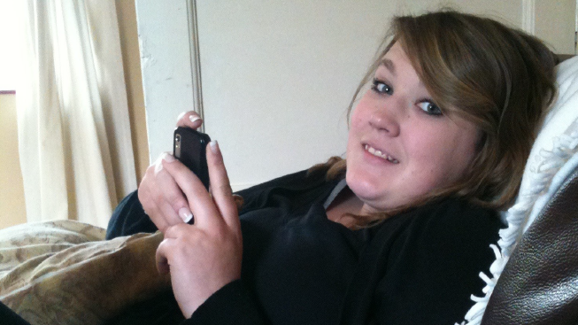 Sophie Alderman, who has shoulder length brown hair and is smiling at the camera. She is lying on a sofa indoors while holding phone in her hands and wearing a black, long-sleeved top.