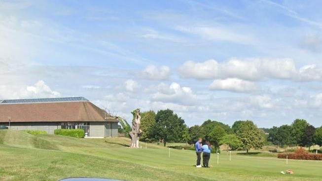 A golf course, with two people dressed in blue and black standing near a hole. There are trees in the background and a clubhouse to the left.