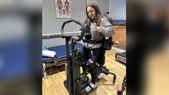 Libby doing physiotherapy using a standing apparatus that is holding her up and she is smiling at the camera with a thumb up.