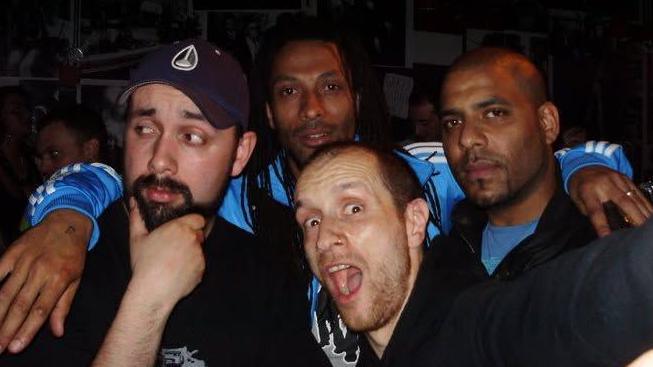 Four people pose closely together for a photograph in a dimly lit indoor venue. One person stands in the foreground at the centre of the group, while three others gather behind and beside them. The image is tightly cropped around the group, with blue sports-style tops visible on two people in the background and dark clothing on others.