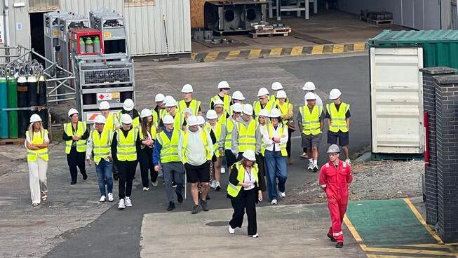 A group of people in yellow high visibility vests are shown around an industrial site, led by a man in a red boilersuit