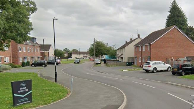 A Google street view image showing a curved, residential street with houses on either side and cars parked in driveways.