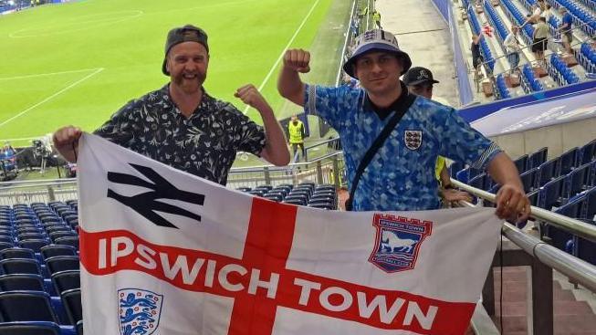 Two men holding an Ipswich Town England flag in an empty stadium with a football pitch in the background