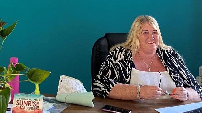 Claire McDowell at her desk speaking to another person. She has blonde hair and is holding her glasses. There is a phone and some flowers on the desk.