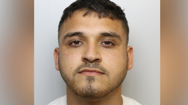 Custody photo of a man with short dark hair, some facial hair and brown eyes. He is wearing a white top and has a scratch on his neck.