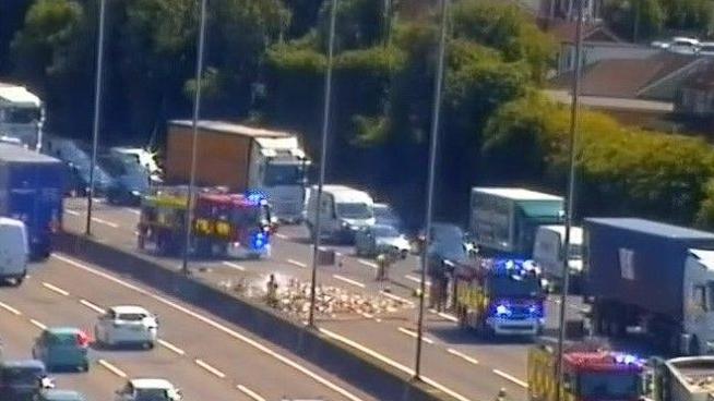 M1 motorway showing queuing traffic on both sides, a fire engines, debris on the road, and vehicles and lorries around it. There are also cars driving on the other side.