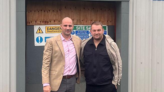 Dale Gaucas wearing a pink shirt and beige blazer and grey pants and Danny Randall with brown hair wearing a black shirt and beige coat and black trousers stand arm in arm in front of the building where they are opening the boxing club. They are smiling.