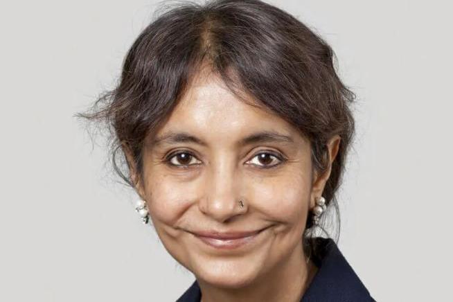 Smiling woman looking at the camera. She is of asian heritage and wearing pearl earrings. The background is a grey colour. 