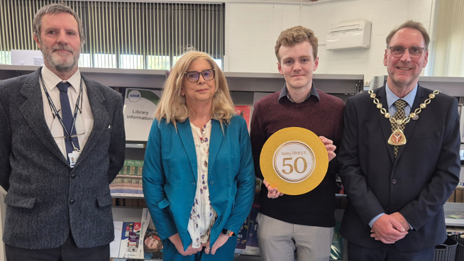 Four people dressed smartly are standing together in front of book shelves in the library.  One man holds an orange circle which reads - Yaxley Library is 50.