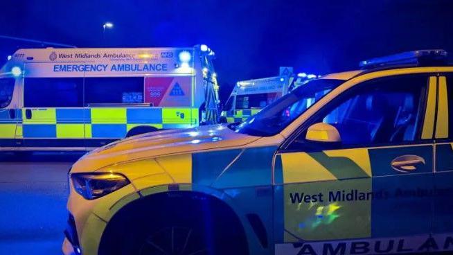 An ambulance service SUV and two ambulance vehicles can be seen at night, with blue lights flashing.