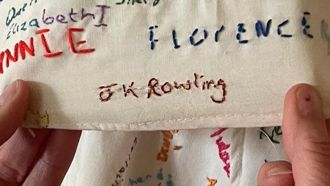 A pair of hands are holding a piece of fabric covered in embroided names. The one being held prominently to the camera is "JK Rowling"