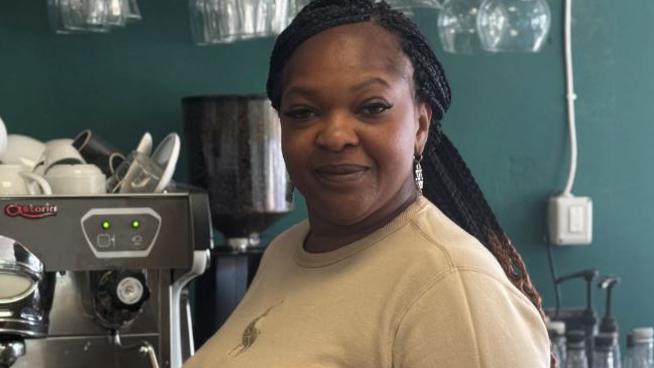 Bukola George, with long dark braided hair, tied back, standing in a cafe by a coffee machine. She is wearing a grey sweater and is looking at the camera and smiling.