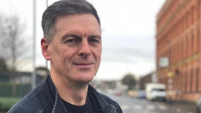 Shaun Harkin standing on a street with buildings, a road and cars behind him. He has black hair and wears a black jacket.