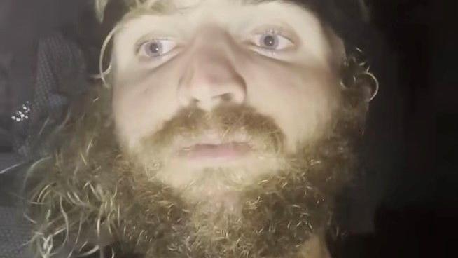 Lachlan has grown a big curly blond beard. It is a close up of his face and darkness surrounds him.