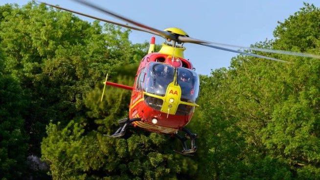 A red air ambulance helicopter, in flight, with trees behind. The helicopter has the letters AA on the front of it.