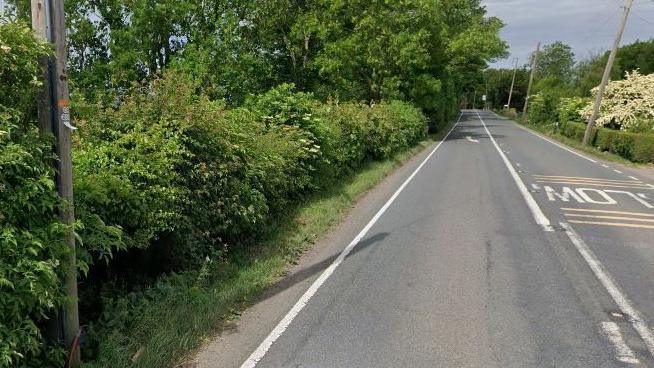 A stretch of an A-road with a hedge on one side and trees in the distance.