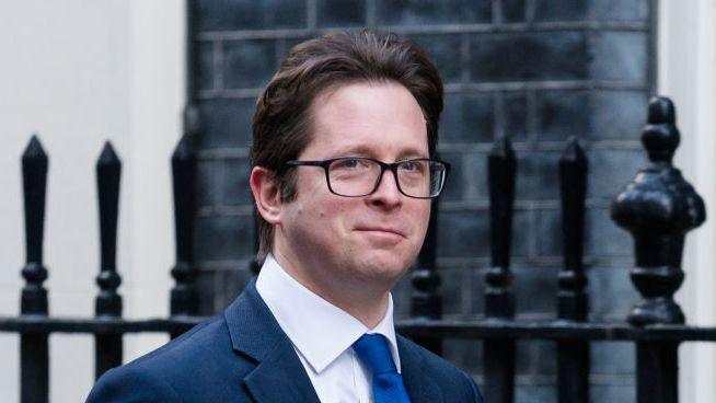 A man with brown hair and dark rimmed glasses is smiling in the direction of the camera. He is wearing a navy blue suit, a white shirt and a bright blue tie. The background is out of focus, but you can see the dark grey brick wall of a building and black metal railings. 