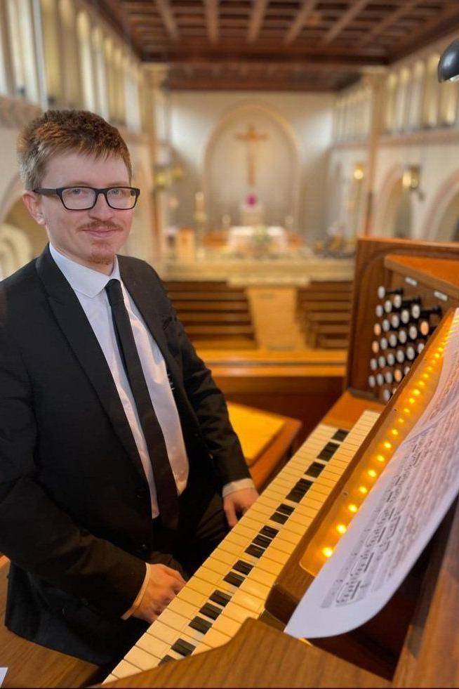 A man with ginger hair and black glasses smiles at the camera, while sat at an organ. He is wearing a black suit and tie with a white shirt. The background of the image, which has been blurred to focus on the man, is that of a large white church with brown palls.