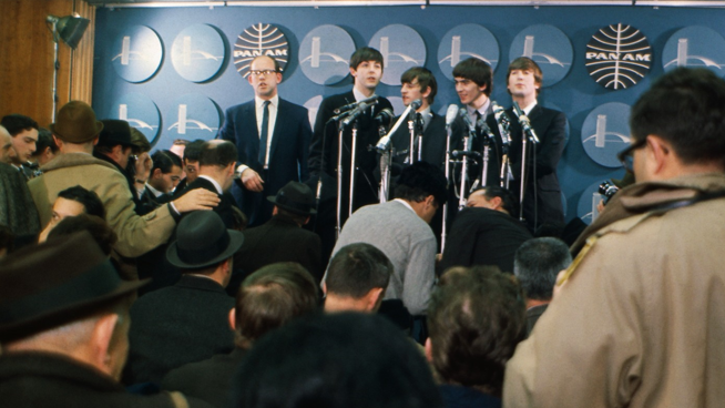 The Beatles stood behind microphones doing a press conference in front of a room full of journalists.