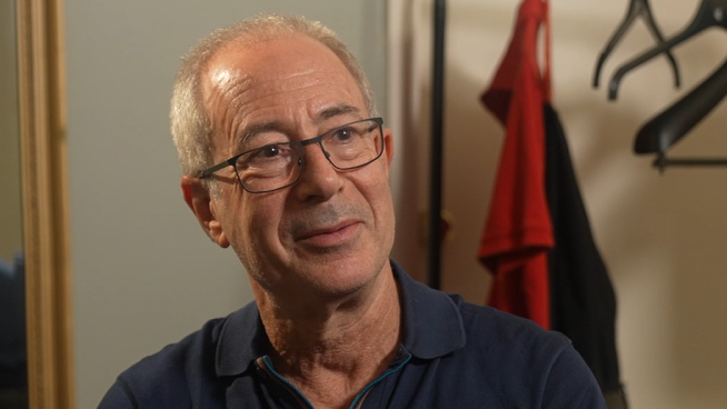The comedian Ben Elton, a man with short grey hair and glasses, wearing a dark blue polo shirt, looks at something behind the camera.