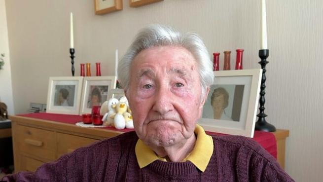 Edwin Rayner has grey hair and is looking directly at the camera. He is wearing a burgundy jumper with a yellow collar. There are images of his late wife Margaret in the background.