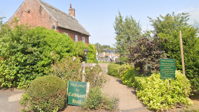 The outside of Coldham Hall in Surlingham. The pub is nestled in shrubbery with green signs out the front which say Welcome to Coldham Hall in gold writing.