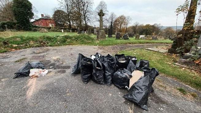 Several black bags filled with rubbish and dumped on a path in a cemetery.