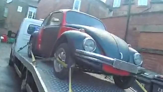 An old-fashioned black and red car, tethered on the back of a delivery truck. Houses are in the background.