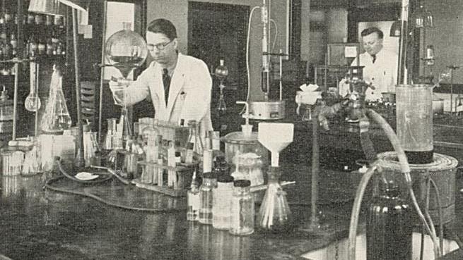 A black and white photo of two men in white lab coats working in a research lab in Leeds. There are lots of flasks and experiments going on on the desks in front of them.
