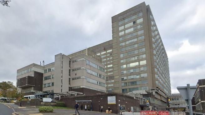 The multi-storey concrete hospital building in Sheffield. Several people can be seen waiting at a bus stop outside the hospital.