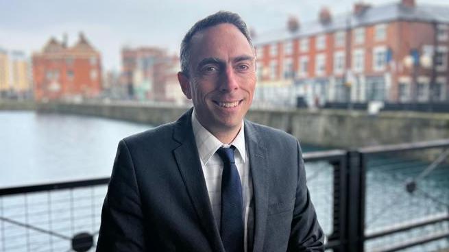 Mike Ross, with short brown hair and wearing a dark suit and navy tie, smiles for the camera against the backdrop of the city's former Princes Quay.