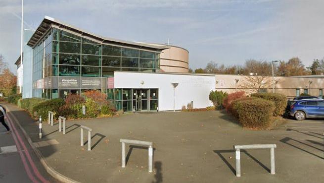 A large building with a glass wall and white entryway. There is a car park on the right hand side