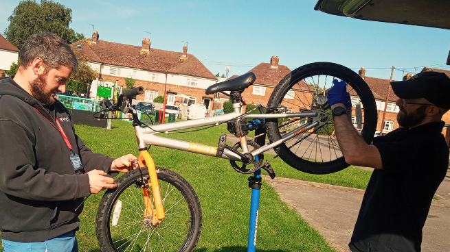 Two men repair a bike near a play park in a residential area with houses in the background. It is sunny. One is working on the front wheel, while another works on the back. The bike is on a clamp.