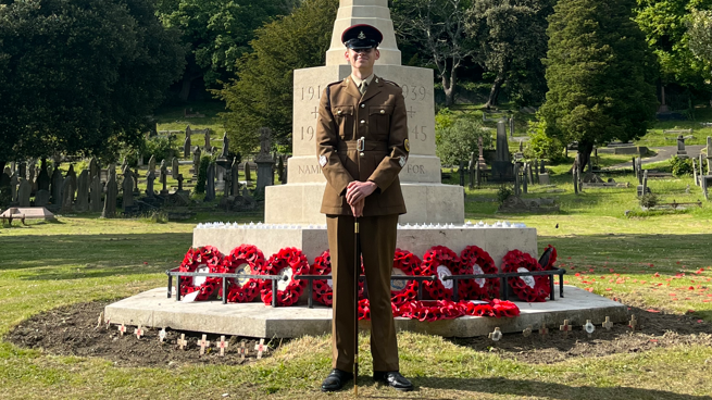 William, a young man in Army cadet uniform standing in front of a war memorial which has several poppy wreaths around the base. He wears a black cap, and leans on a cane. 