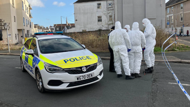 A police cordon in Plymouth. There are four forensics officers and a police officer huddled on the right of a police car positioned in the road, behind police tape.