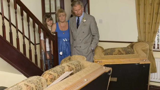 A blonde woman stands with a younger girl next to a younger King Charles as they look down at a flooded lounge with an upturned sofa.