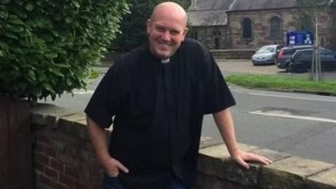Rev Huw Thomas in clerical attire leaning on a wall near his church.