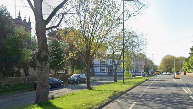 A street view of a road with trees on a grassy verge and a church to the left with a black and white Tudor-esque pub behind it.