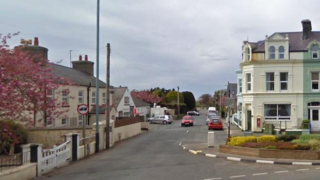 The start of the A10 road at Ballaugh Bridge. there are buildings on both sides, a traffic island with planters on it to the right and a blossoming cherry tree to the left. There are parked and moving cars in the carriageway.