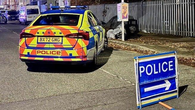 A police car in the middle of a road at night with a diversion sign placed in the way of traffic