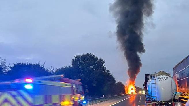 A fire on the M5 with smoke billowing from above which is dark and grey. There is a fire service in the corner with flashing lights. 