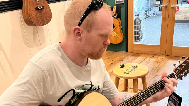 Craig Brown, seated, playing the guitar in a shop with guitars mounted on the wall behind him