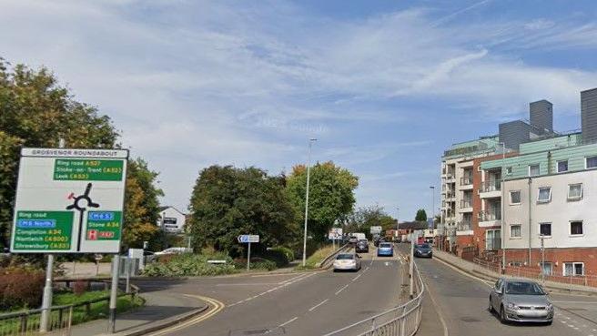 A dual carriageway with a large road sign on the left hand side, a building on the right hand side and a central reservation. There are trees to the left further into the distance.