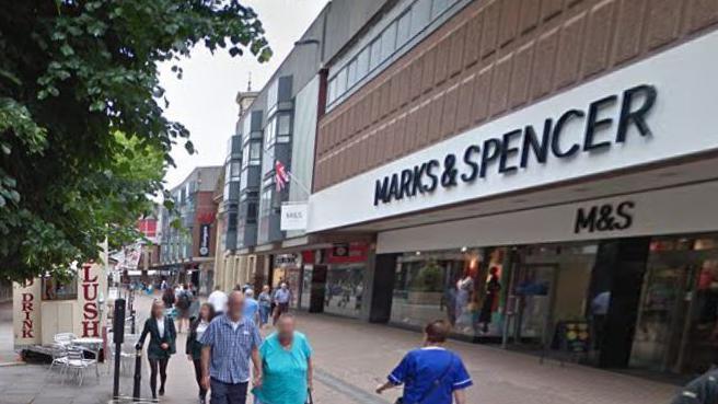 Entrance to Marks and Spencer shop in Gloucester. Several shoppers walking past including a woman in nurse's outfit and an elderly couple holding hands.